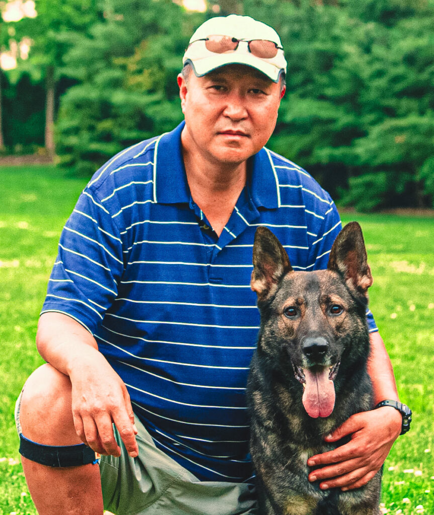 Ki Hong, wearing a striped blue polo shirt and sunglasses perched atop his cap, is squatting on a lush green field. He gently rests his hand on the shoulder of his attentive German Shepherd, identified as Kahn's father, who looks directly at the camera with a tongue out, suggesting a warm and tranquil day outdoors.