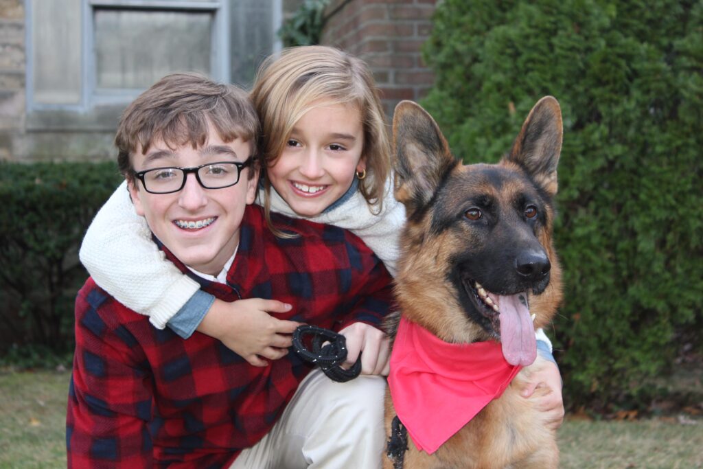 Finley, a happy German Shepherd wearing a red bandana, poses with a smiling boy and girl, both in festive plaid attire, showcasing a warm family moment