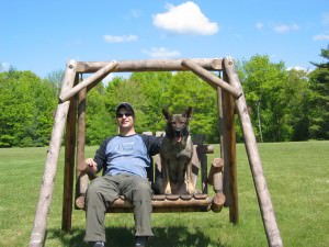 A smiling dog owner and Kara, a German Shepherd, seated side by side on a rustic wooden swing in a sunny park, exuding a sense of accomplishment and companionship.